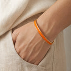 Orange enamel reminder cuff worn as one continuous band with a hand resting in a linen pocket