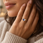 Two-birthstone heart ring with blue and clear stones in a gold finish, worn on a hand beside the face