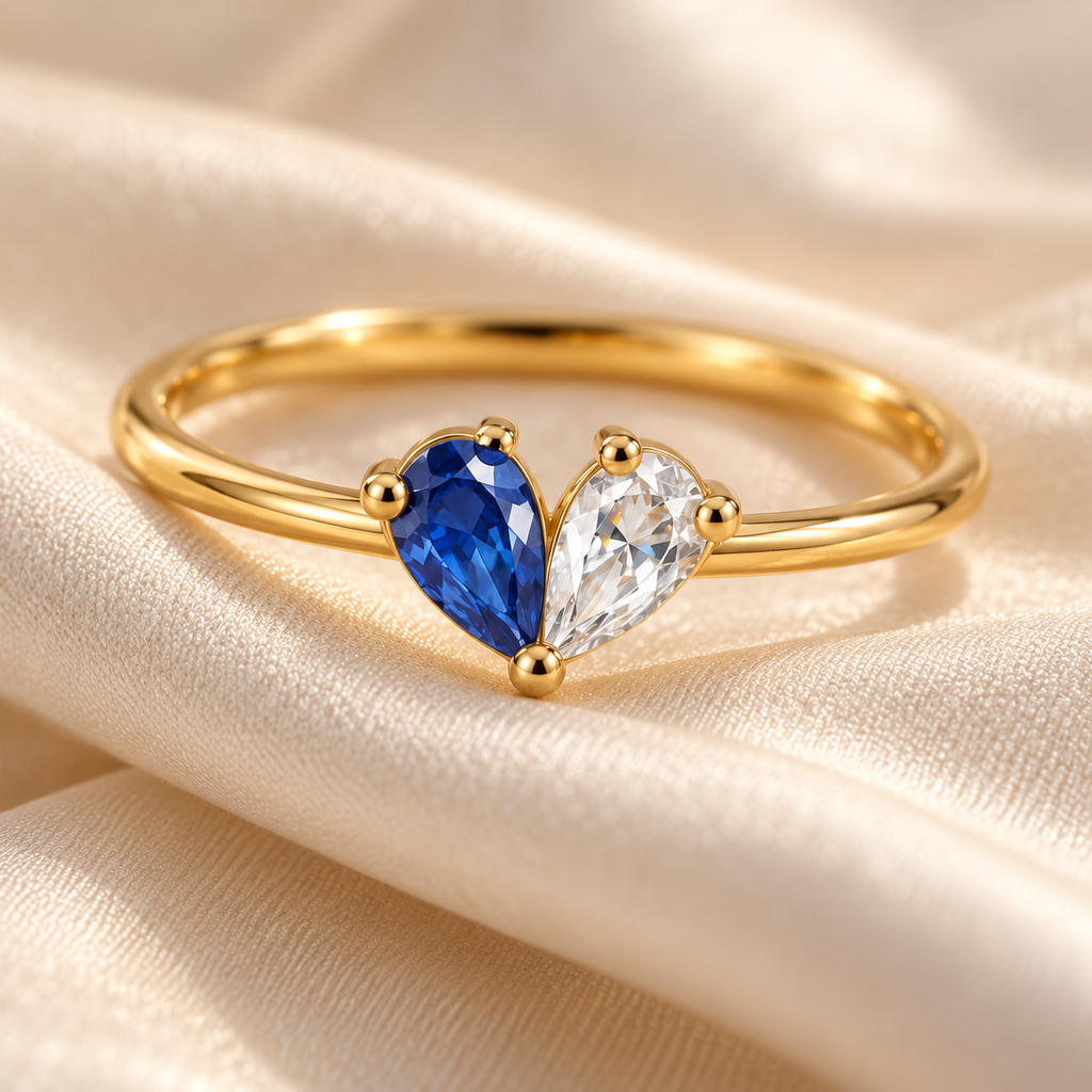 Close-up of the gold-finish two-birthstone heart ring with blue and clear pear-cut stones on cream fabric