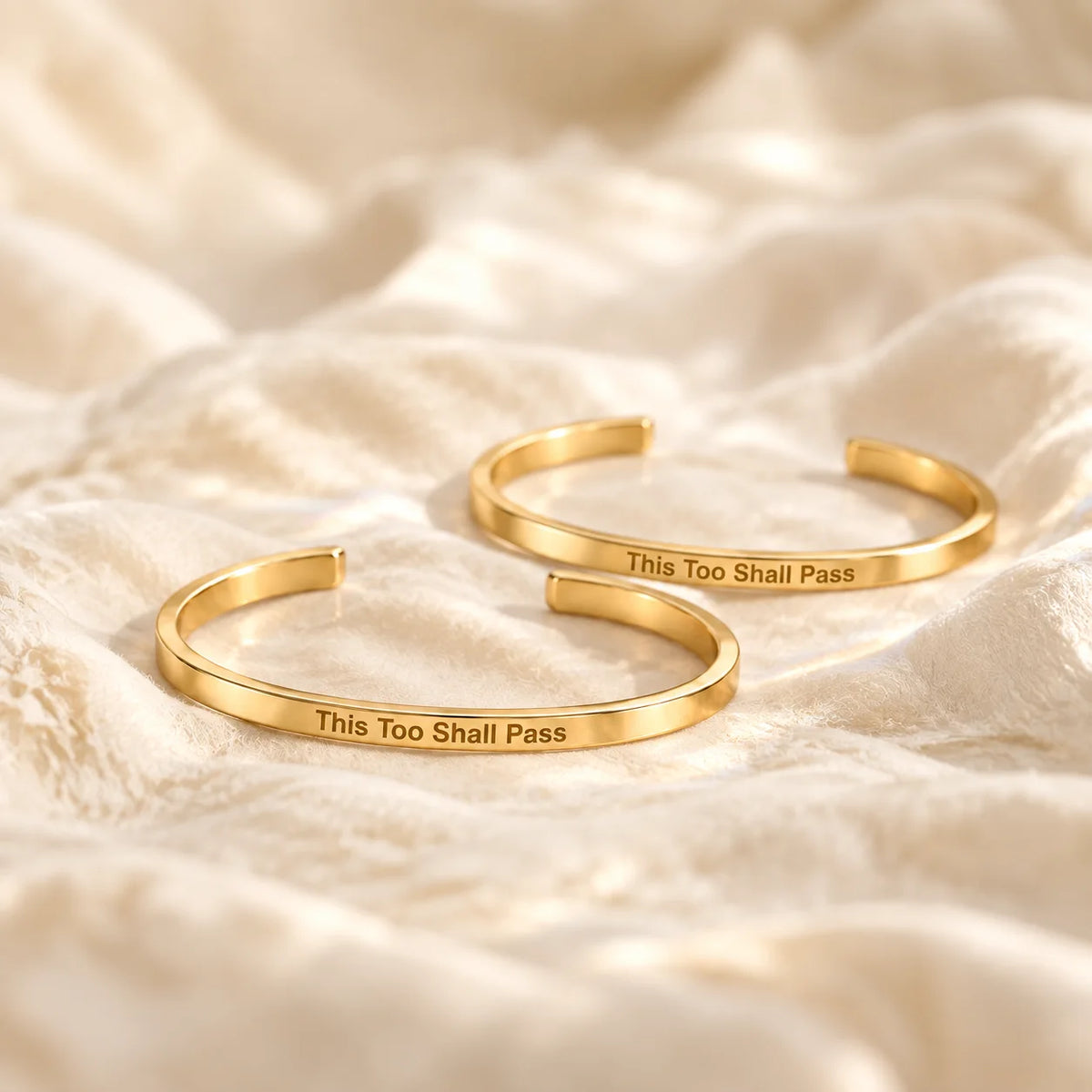 Two gold This Too Shall Pass Slim Cuffs with outside engraving on ivory fabric